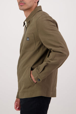 A man stands sideways against a plain light background, one hand in his pocket, wearing the olive green Swanndri Northgate Canvas Jacket featuring a chest pocket and trucker style design.