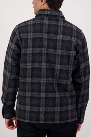 A person is shown from behind wearing the Swanndri Northgate V2 Jacket in a dark plaid pattern with gray stripes over black pants, standing against a plain light background.