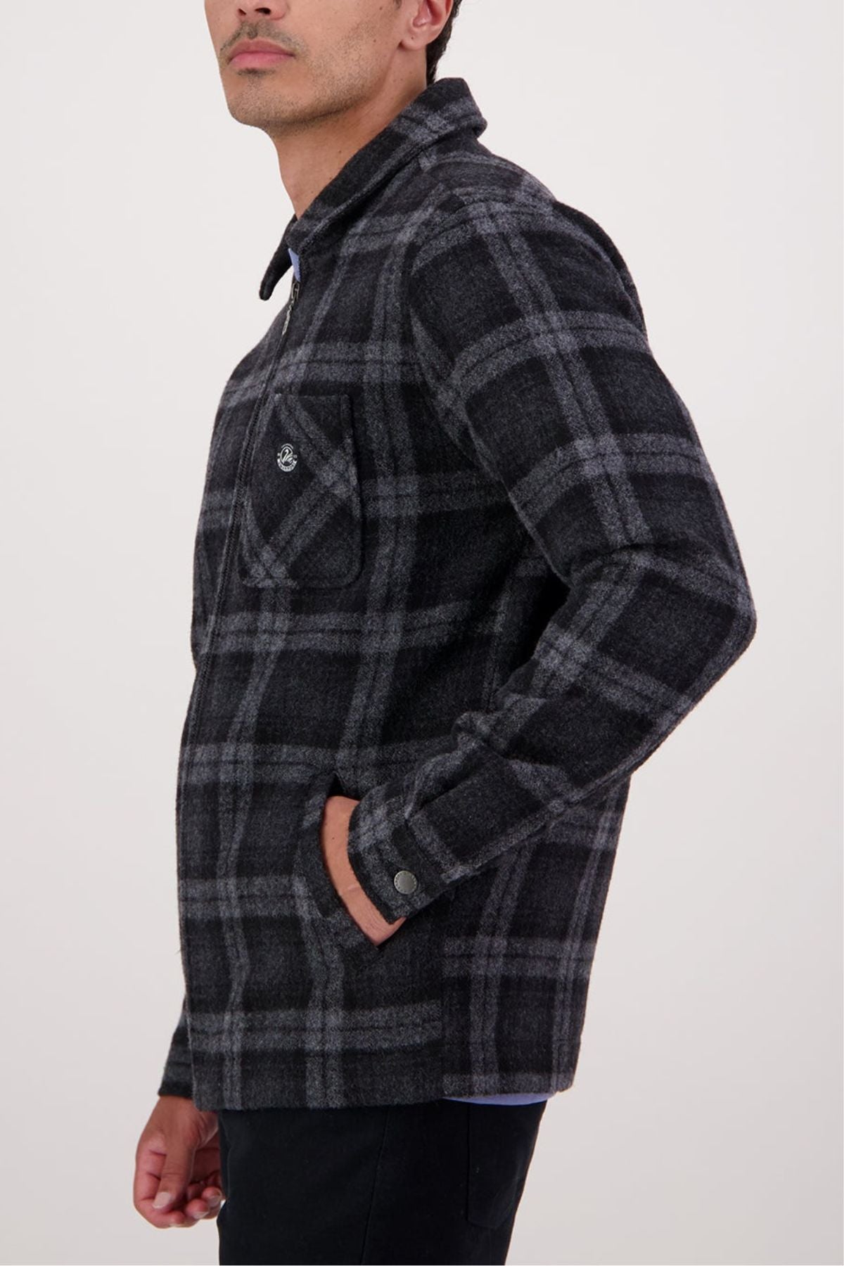 A person stands sideways with one hand in their pocket, wearing the Swanndri Northgate V2 Jacket in a black and gray plaid pattern. The jacket features a chest pocket and is set against a plain light background.