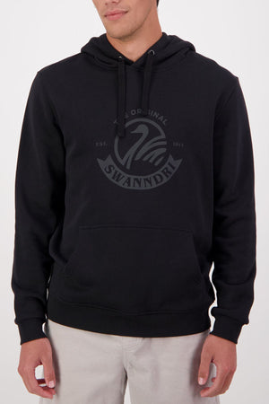 Someone wears the Swanndri Original V2 Hoodie—a black cotton-polyester hoodie featuring a kangaroo pocket and a subtle Swanndri logo with swan graphic on the chest—paired with light-colored pants.