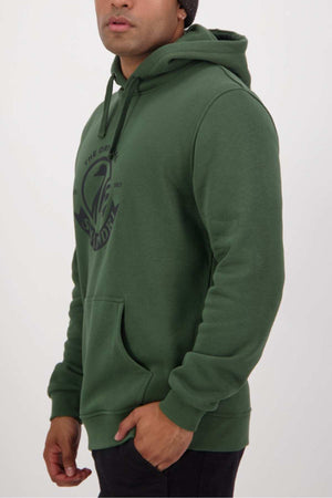 Clad in a green Swanndri Original V2 Hoodie, which boasts a circular black logo with graphic and text on the chest, the person stands with their hands by their sides, facing slightly to the left. The cotton and polyester blend hoodie vividly contrasts against the plain, light backdrop.