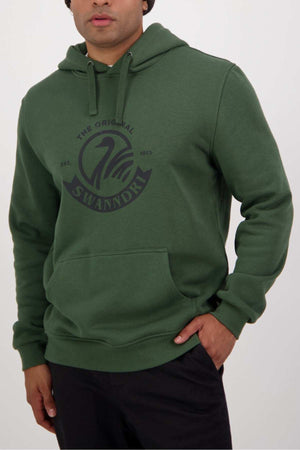 A person is wearing a green Swanndri Original V2 Hoodie, featuring "The Original Swanndri Est. 1913" logo in dark text on the front. The hoodie, made from a cotton and polyester blend, includes a kangaroo pocket and drawstrings at the hood. They are also dressed in black pants, with their left hand partially tucked into the pocket.