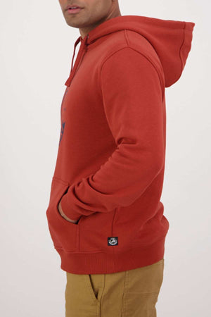 A person wearing a red Swanndri Original V2 Hoodie with their hands in the front pocket and tan pants is shown from the side against a plain background.