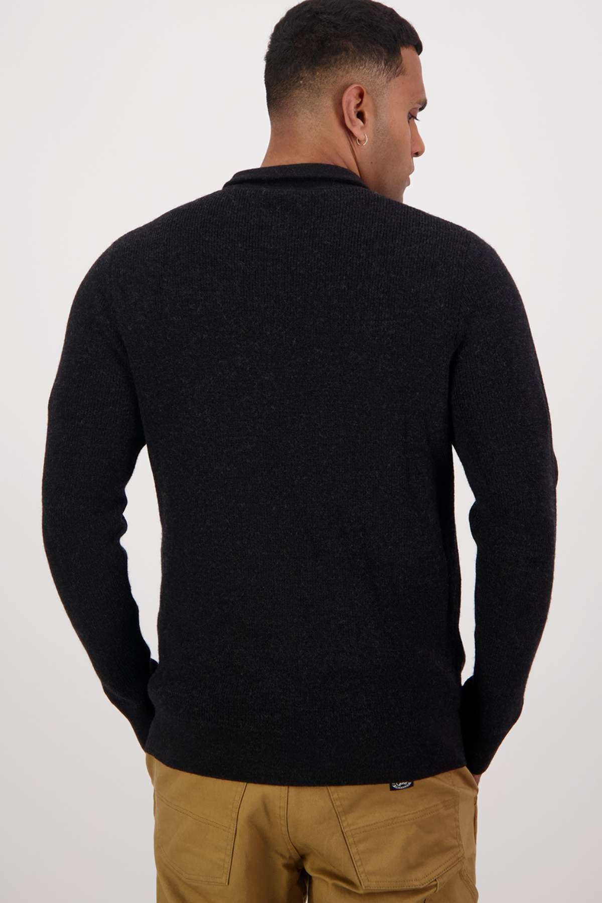 A person with short hair, wearing a Swanndri Otahu River Quarter Zip Merino Knit sweater and tan pants, faces away. The plain, neutral background highlights their clothing.