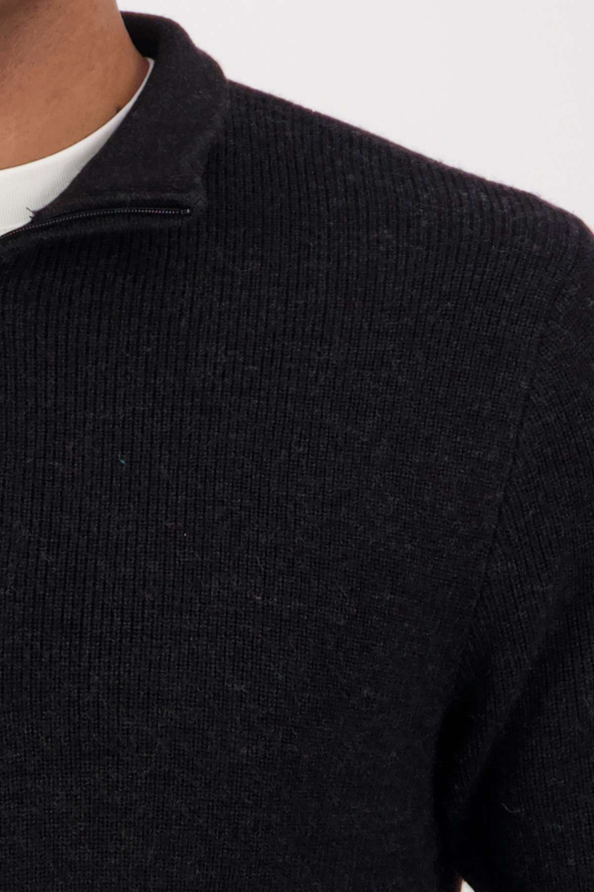 A close-up shows a person wearing the Swanndri Otahu River Quarter Zip Merino Knit sweater, featuring ribbed, dark merino with a quarter zip near the collar. It's layered over a white shirt against a plain, light-colored background.