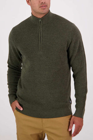 A man in an olive green Swanndri Otahu River Quarter Zip Merino Knit sweater and tan pants stands against a plain light background with one hand in his pocket.