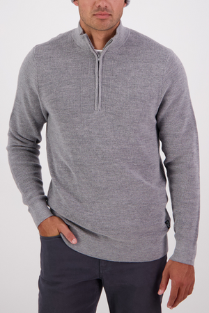 A man wears the Swanndri Otahu River Quarter Zip Merino Knit sweater in light gray with dark gray pants, standing with one hand in his pocket against a plain white background.