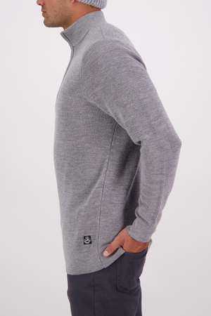 A man in dark pants wears the Swanndri Otahu River Quarter Zip Merino Knit sweater in grey, featuring a high collar, quarter zip, and small black hem logo. He stands in profile with one hand in his pocket against a plain white background.