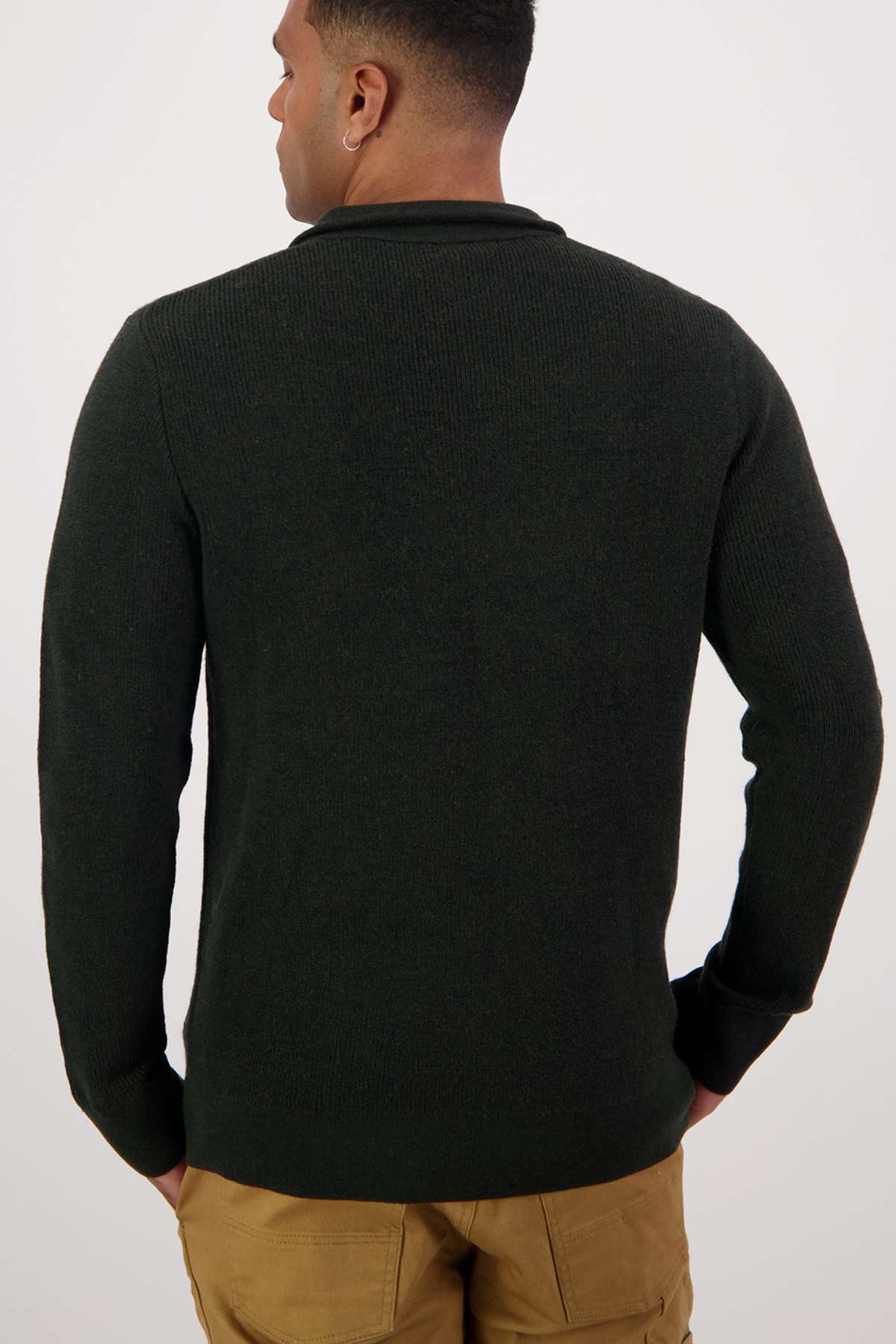 A man faces away from the camera, wearing a Swanndri Otahu River Quarter Zip Merino Knit sweater in dark color and tan pants, his hands at his sides against a plain light background.