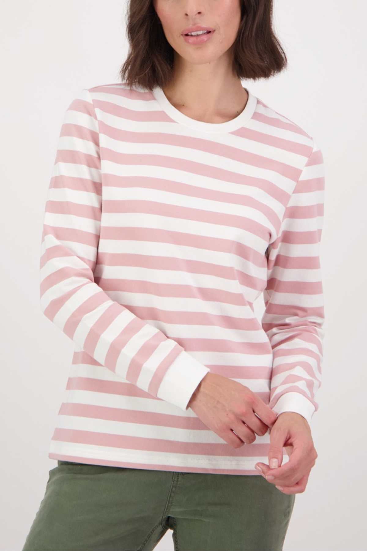 A woman models the Swanndri Papamoa Stripe Long Sleeve Tee in pink and white with white cuffs and a relaxed fit, paired with olive green pants. She stands against a plain background, her brown hair framing her face—a versatile look for everyday wear.