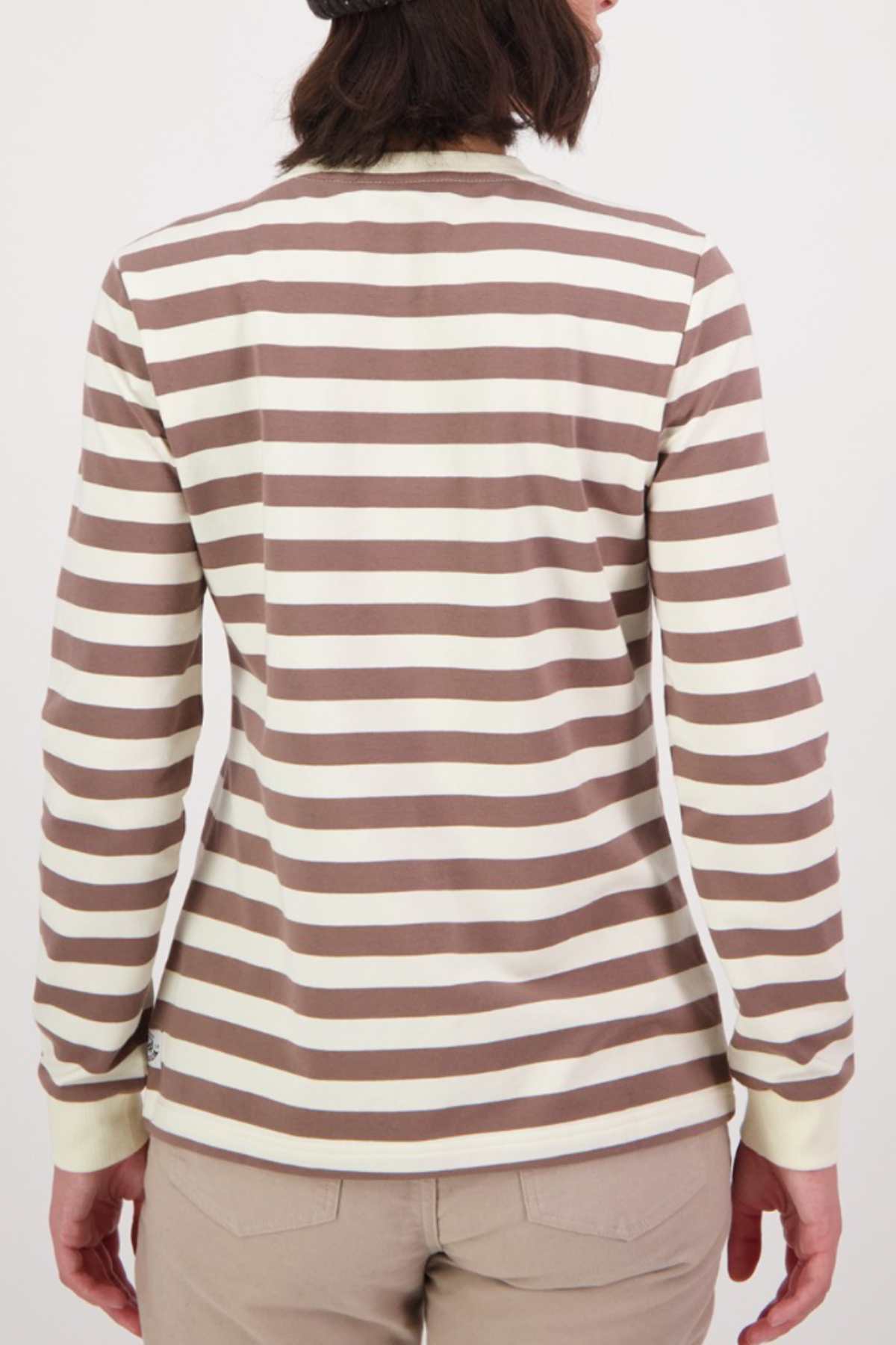 A person with shoulder-length dark hair wears the Swanndri Papamoa Stripe Long Sleeve Tee, made from cotton elastane, showcasing white and brown stripes. Their light brown pants stand out against a plain white background.