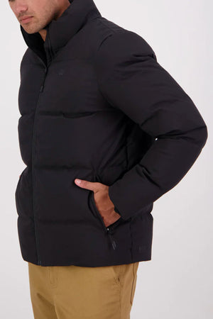 A person stands sideways on a plain white background, wearing light brown pants and a black Swanndri Plume Mens Down Jacket filled with duck down for warmth. Their left hand is in the pocket, making this jacket perfect for outdoor activities.
