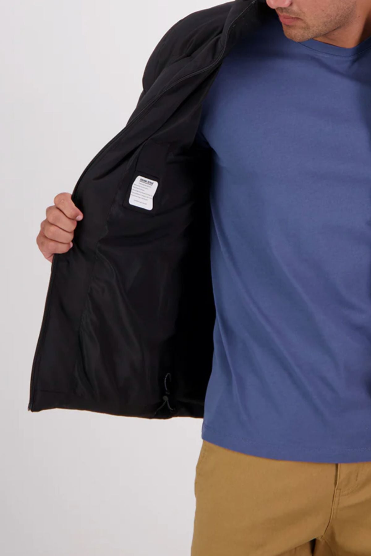 A person in a blue shirt and tan pants holds open a Swanndri Plume Mens Down Jacket, revealing its interior lining and white tag. The person's face is partially out of frame. This jacket is insulated with Duck Down, making it perfect for outdoor use.