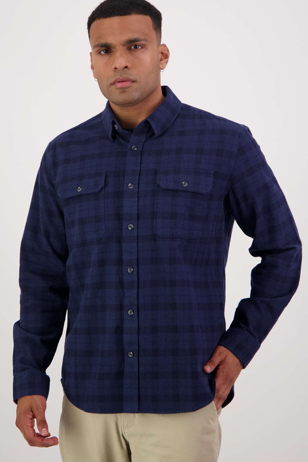 A man stands against a plain light background wearing the Swanndri Port Road Shirt, which features a navy blue and black plaid heavyweight twill design. He has short hair, a neutral expression, and one hand in his pocket.