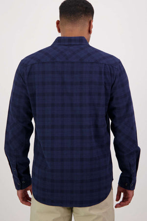 A man is seen from behind wearing the Swanndri Port Road Shirt, a dark blue plaid heavyweight twill long-sleeve shirt, paired with light-colored pants against a plain light background.