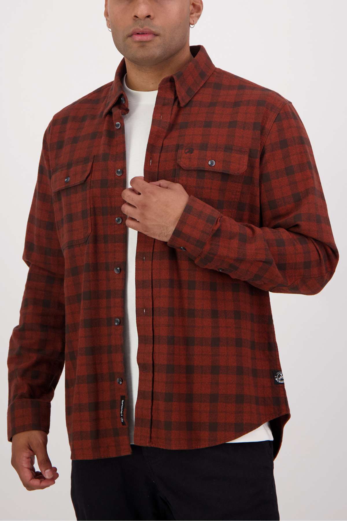 Wearing a Swanndri Port Road Shirt, this individual artfully adjusts the rust-colored plaid shirt with buttoned chest pockets over a white t-shirt. The cotton shirt pairs seamlessly with black pants against a plain white background.