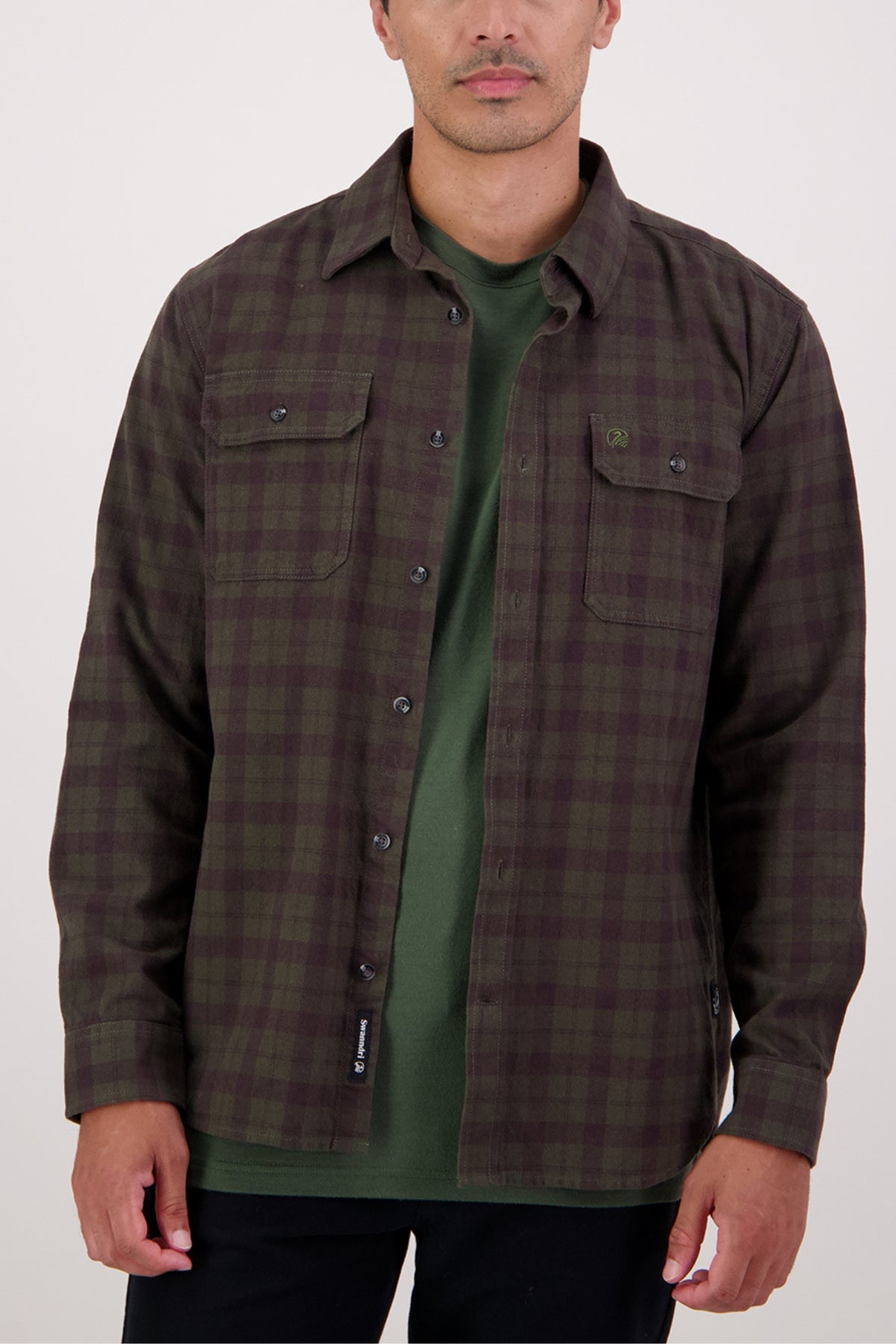 A man wears a dark green and brown plaid Swanndri Port Road Shirt over a green t-shirt, standing against a plain light background. This heavyweight twill shirt features two chest pockets and black buttons.