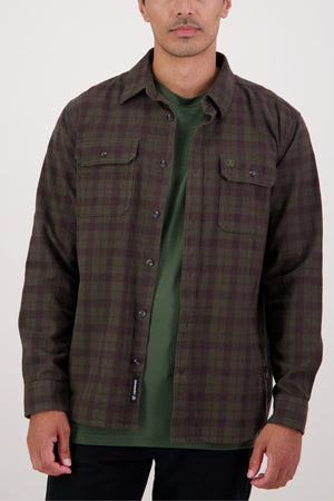A man wears a dark green and brown plaid Swanndri Port Road Shirt over a green t-shirt, standing against a plain light background. This heavyweight twill shirt features two chest pockets and black buttons.