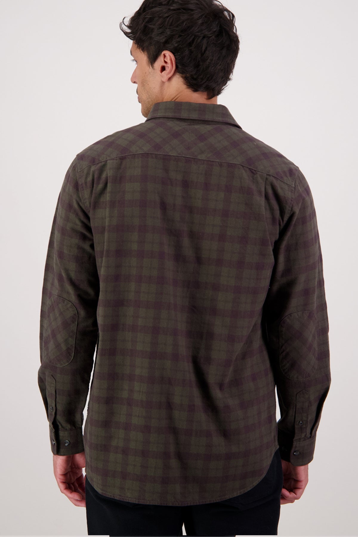 A man with dark hair stands with his back to the camera, wearing a Swanndri Port Road Shirt in dark green and brown plaid and black pants against a plain light background.