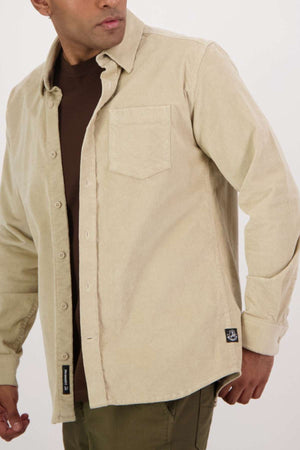 A person wears the Swanndri Ranfurly Corduroy Shirt—a heavy, tailored beige cotton shirt with a chest pocket and black label—over a brown tee and olive green pants, standing against a plain light background.
