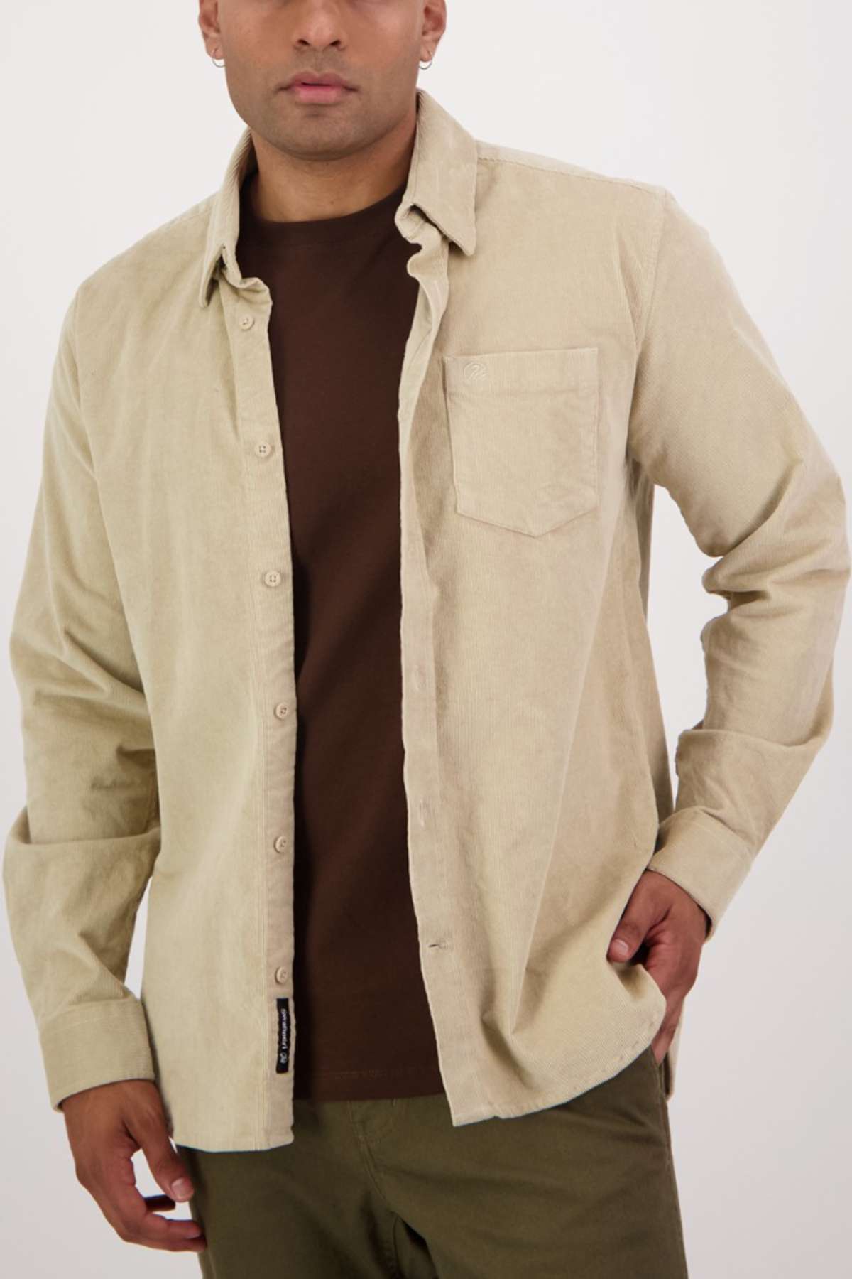 A person with short hair is wearing a light beige, tailored-fit Swanndri Ranfurly Corduroy Shirt over a dark brown T-shirt. The heavy weight cotton shirt is unbuttoned, has long sleeves, and features a pocket on the left side. The person is posing with one hand in their pocket against a plain background.