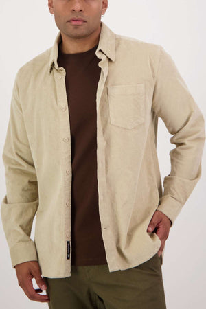 A person with short hair is wearing a light beige, tailored-fit Swanndri Ranfurly Corduroy Shirt over a dark brown T-shirt. The heavy weight cotton shirt is unbuttoned, has long sleeves, and features a pocket on the left side. The person is posing with one hand in their pocket against a plain background.