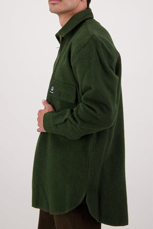 A person wearing an oversized Swanndri Ranger Bush Shirt with a front pocket stands against a plain light background, shown from shoulders to mid-thigh, facing sideways with their hand near the pocket.