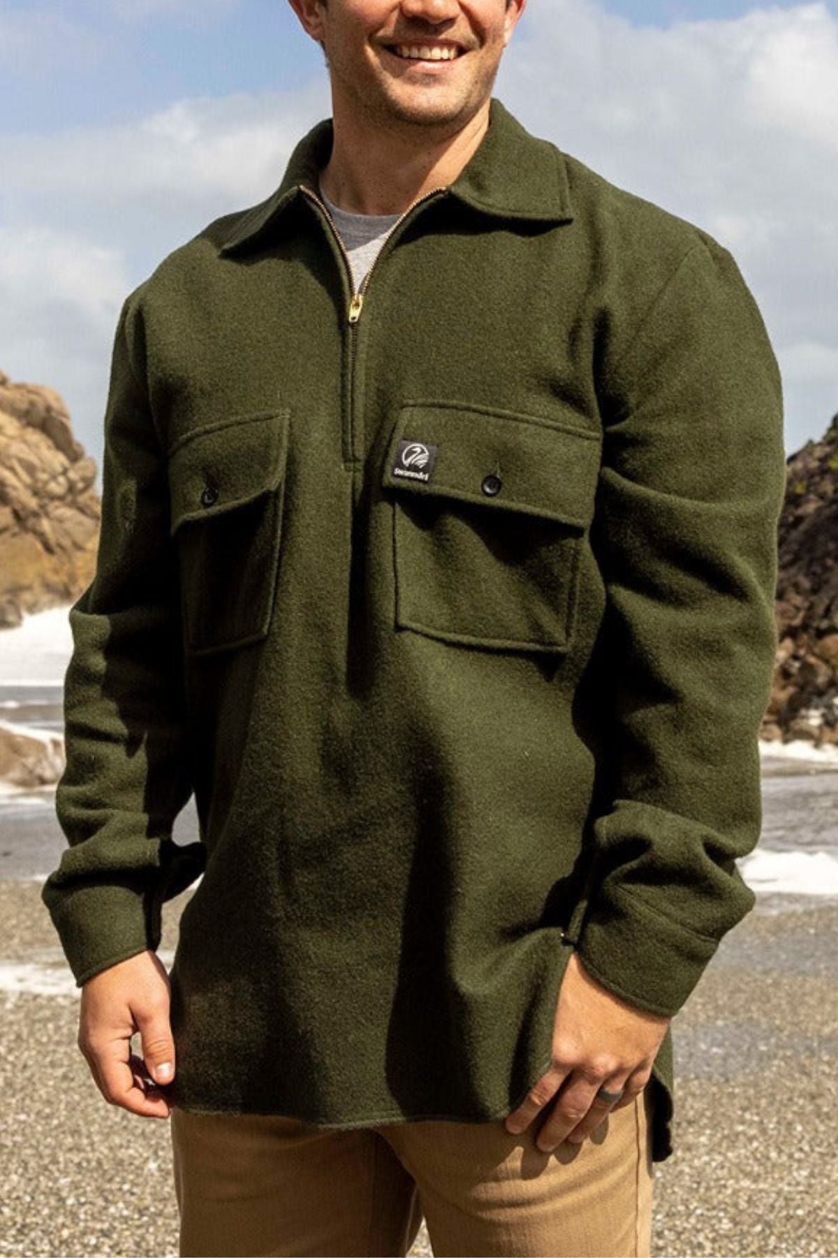 A man wearing a Swanndri Ranger Bush Shirt in olive green with large chest pockets stands on a rocky beach, with the ocean and sky behind him.