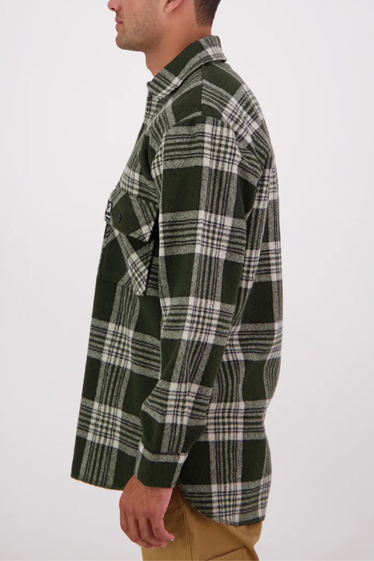 A person stands sideways in a Swanndri Ranger Bush Shirt—green and white plaid, oversized fit with front pocket—paired with light brown pants, against a plain white background.