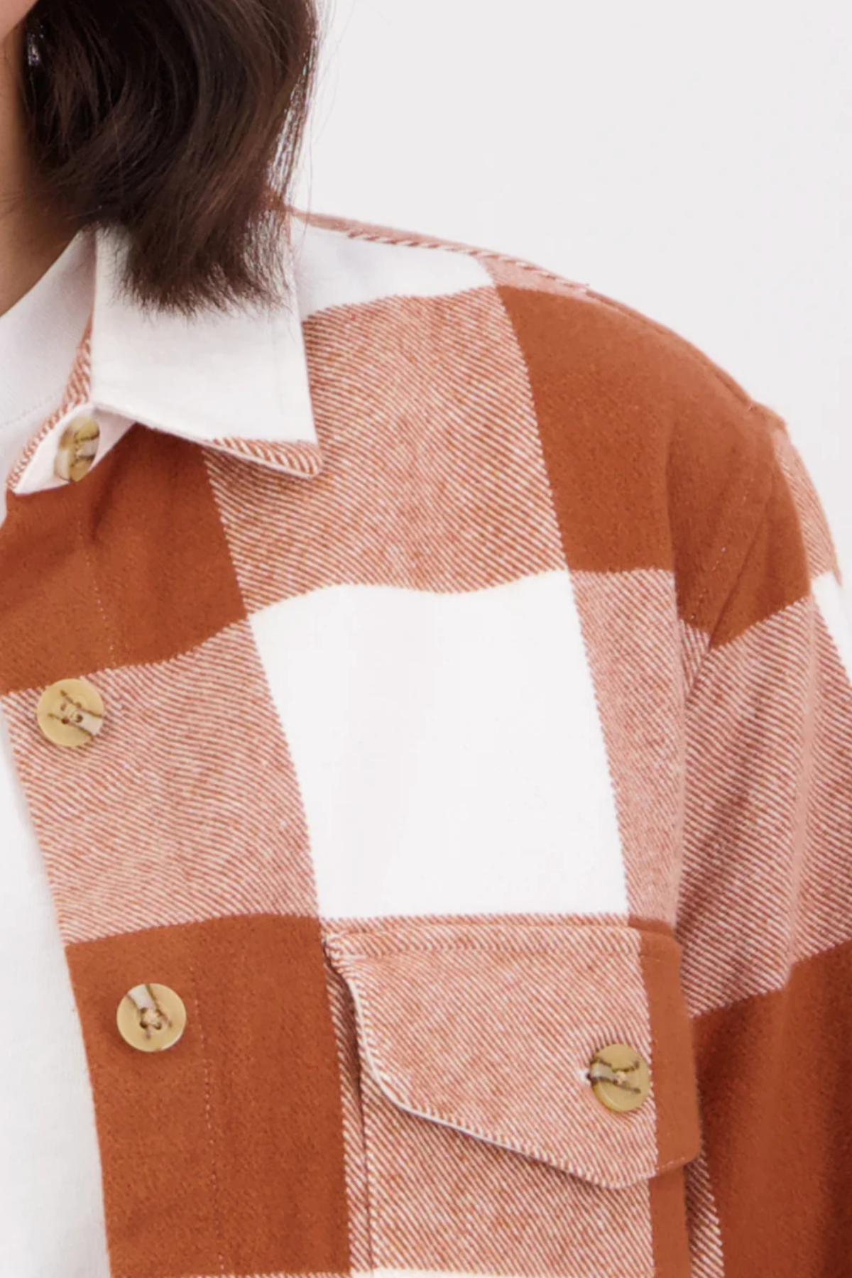 Close-up of a person wearing the Swanndri Rapaki Long Sleeve Shacket, made from 100% brushed cotton in rust and white plaid with large buttons and a front pocket—ideal as a winter layer over a white undershirt.