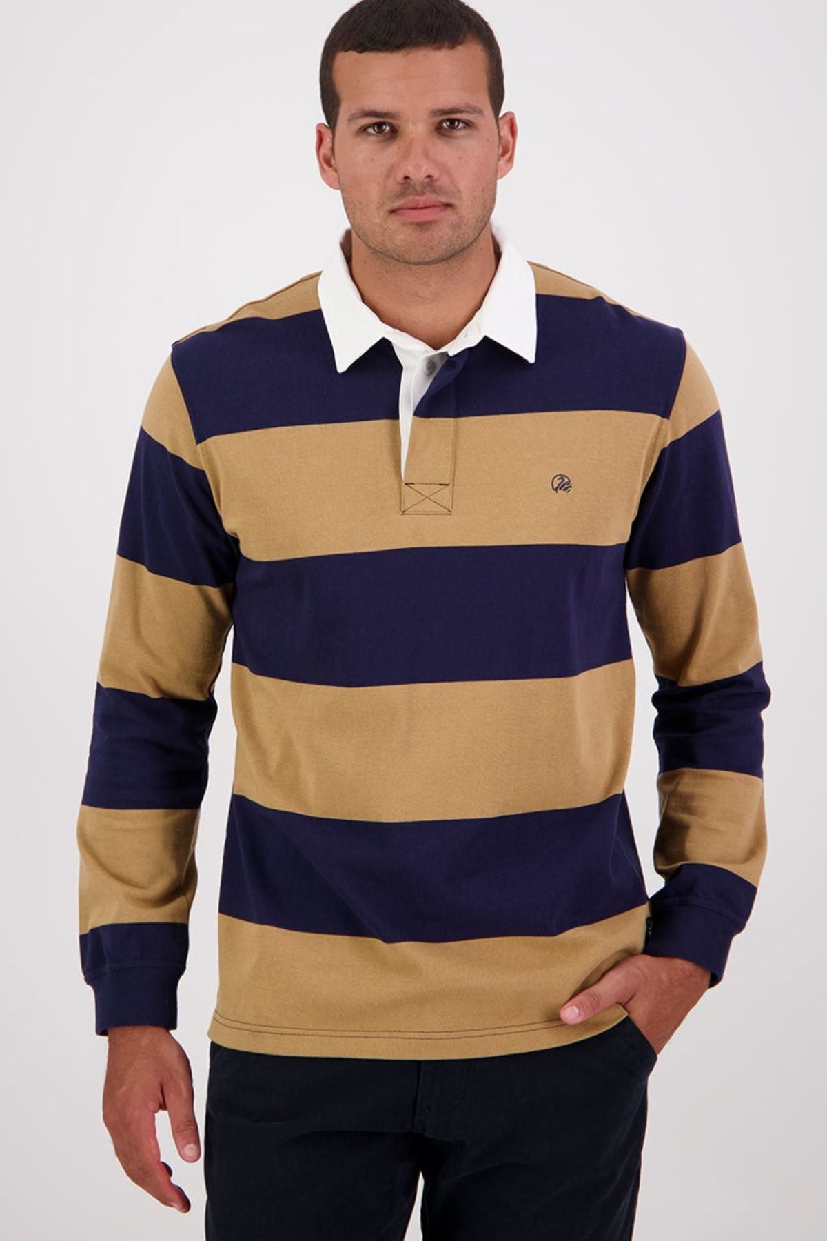 A man faces forward wearing the Swanndri Ridgeway Rugby, a long-sleeve cotton shirt with wide navy blue and tan stripes, rubber buttons, a white collar, and dark pants against a plain light background.