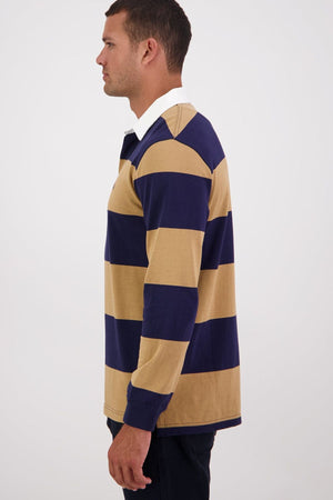 A man in profile wears the Swanndri Ridgeway Rugby, a cotton top with wide navy blue and tan stripes, rubber buttons, and a white collar, standing against a plain light background.