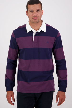 A man wearing the Swanndri Ridgeway Rugby—a long-sleeve shirt in heavy drill cotton with wide purple and navy stripes, a white collar, and rubber buttons—stands facing forward against a plain light background.