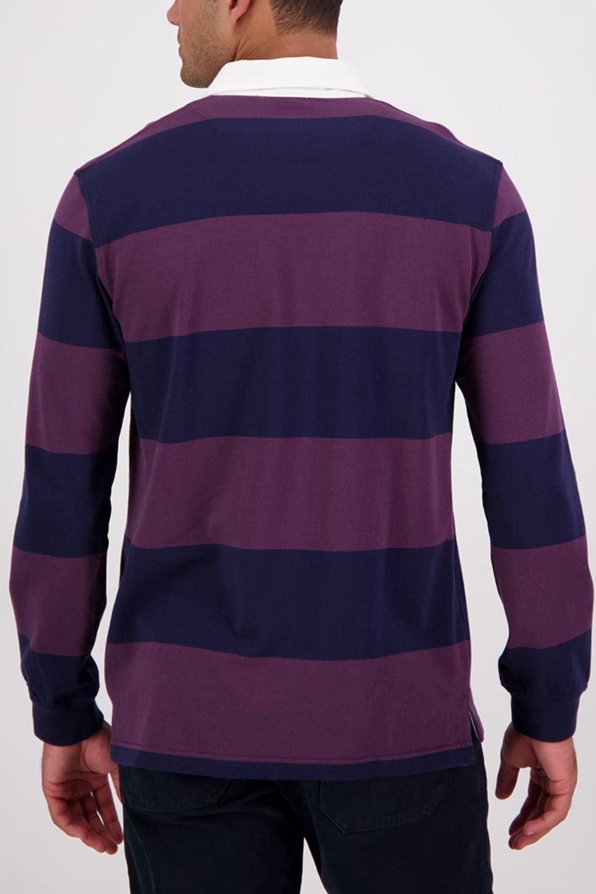 A man is seen from behind wearing the Swanndri Ridgeway Rugby, a long-sleeve collared shirt with wide navy and purple stripes, rubber buttons, and crafted from heavy drill cotton, standing against a plain light background.