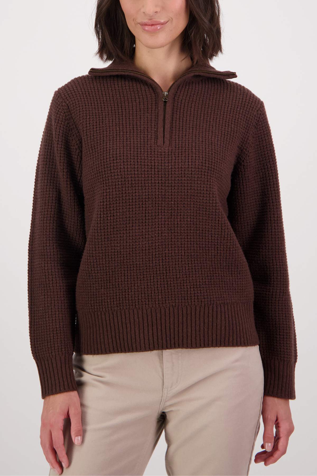 A person wears the Swanndri Riverside v2 Zip Neck Rib Jumper in brown, featuring a half-zip collar and textured knit, paired with light beige pants, while standing against a plain white background.