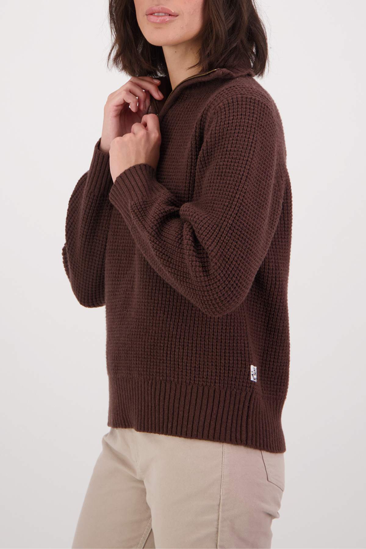 A woman with shoulder-length brown hair wearing the Swanndri Riverside v2 Zip Neck Rib Jumper in dark brown and light beige pants stands against a plain white background, touching her sweater collar and facing slightly sideways.