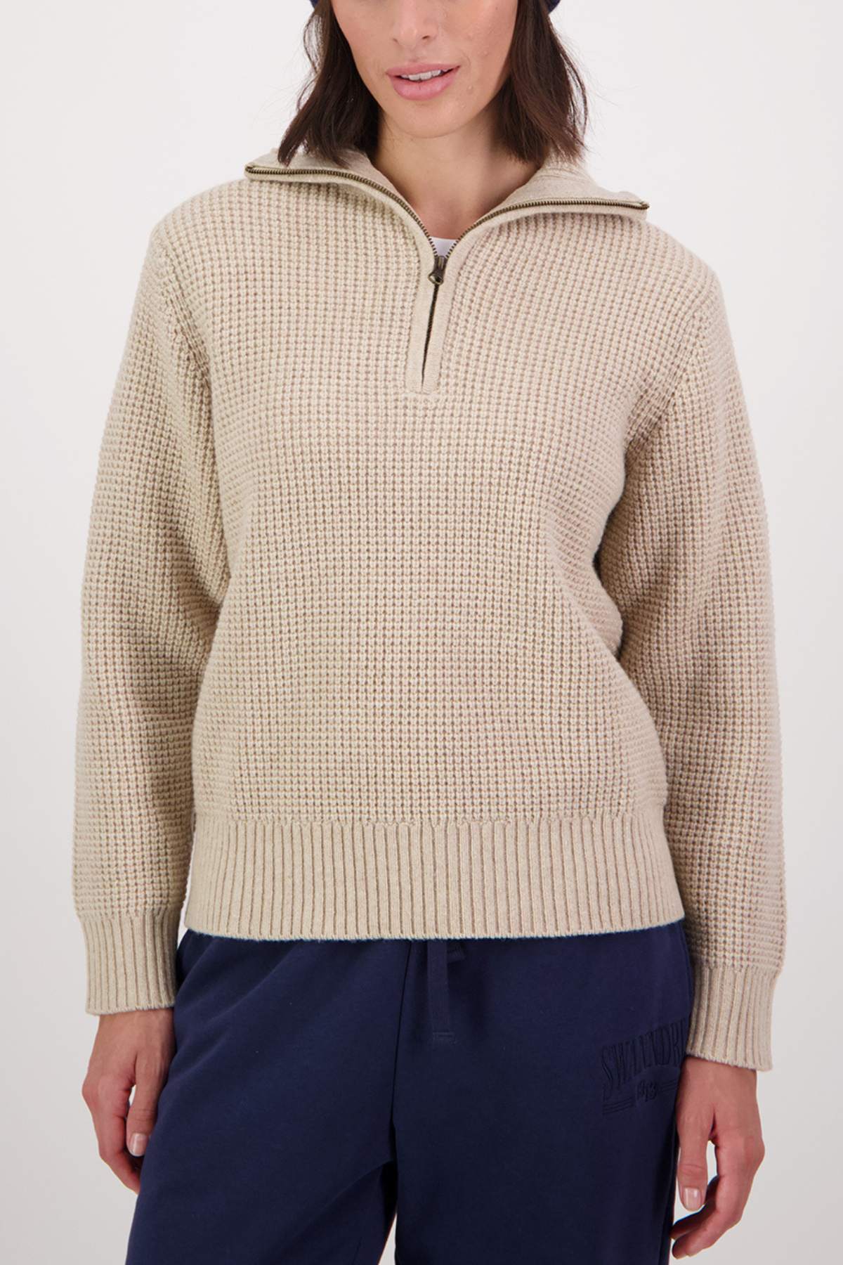 A woman wears a beige Swanndri Riverside v2 Zip Neck Rib Jumper with a collar, paired with dark blue pants, standing against a plain light background.