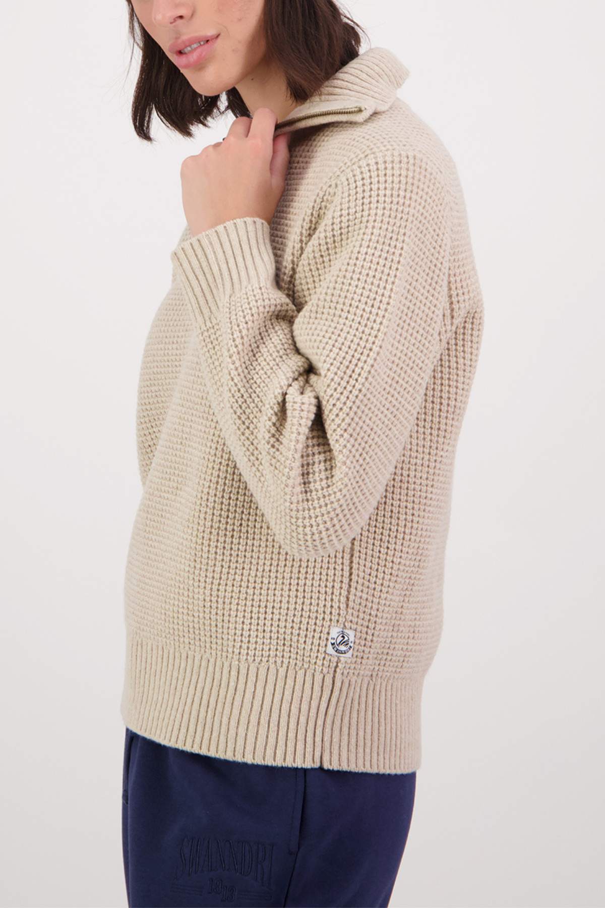 A person wearing a cream-colored Swanndri Riverside v2 Zip Neck Rib Jumper holds the collar with one hand. Paired stylishly with dark blue pants, the image is cropped around the shoulders and knees against a plain white background.