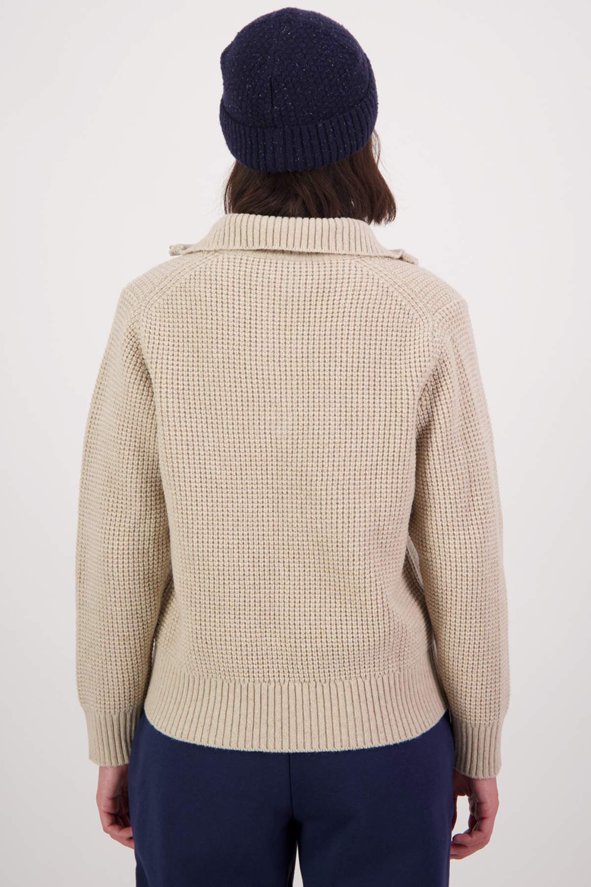 Rear view of a person in a cozy Swanndri Riverside v2 Zip Neck Rib Jumper and dark beanie against a plain backdrop. They have shoulder-length hair and are wearing dark pants.