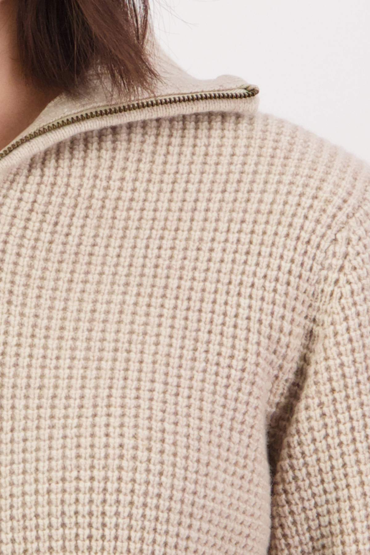 A close-up of a person wearing the Swanndri Riverside v2 Zip Neck Rib Jumper, featuring a beige waffle-knit texture with a zipper detail at the collar. The plain white background accentuates the cozy elegance of this timeless piece, highlighting the shoulder and neck area.