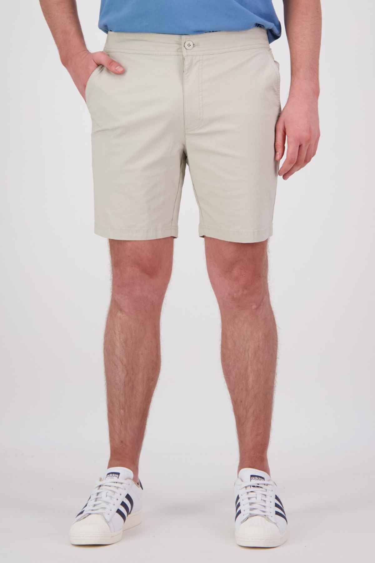 A person wearing Swanndri Sandy Point Shorts in a light beige color made from stretch cotton fabric, paired with a blue shirt and white sneakers featuring black stripes, stands with their hands in pockets against a plain background.