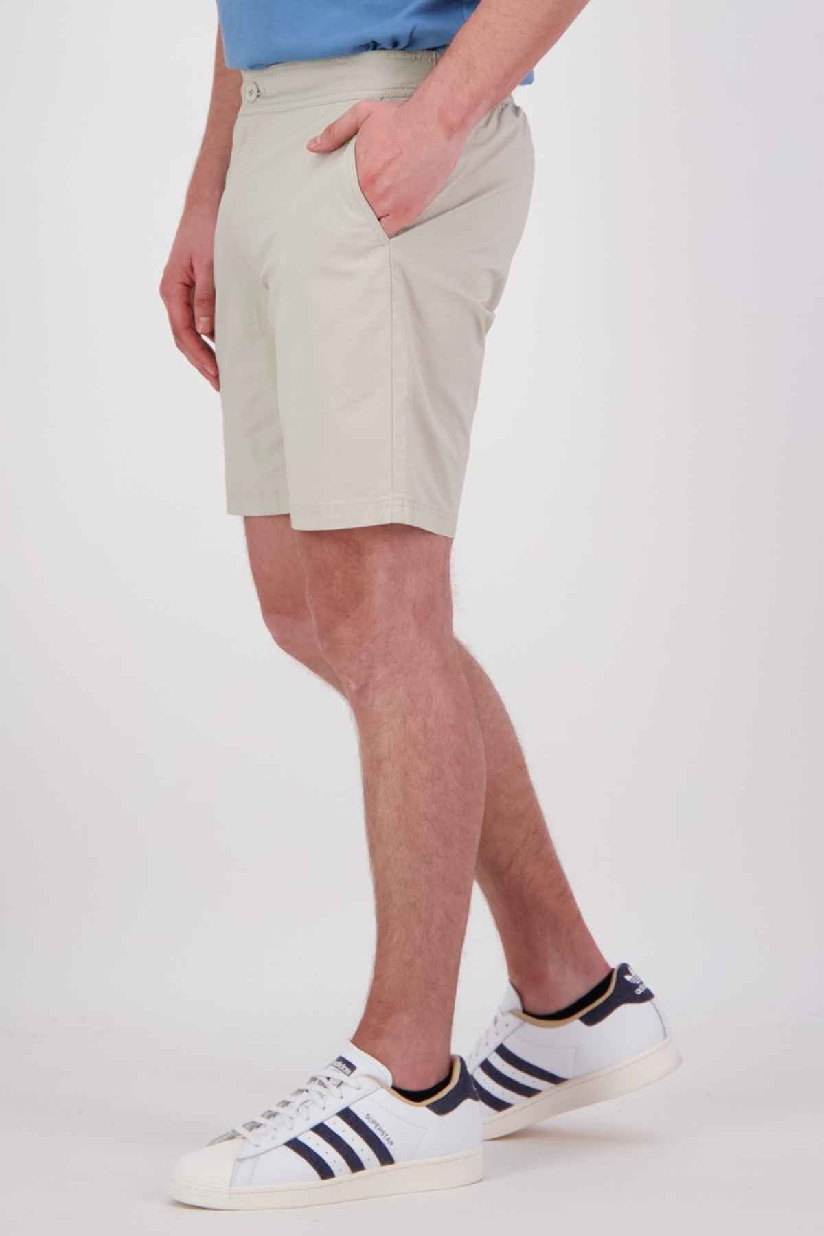 Against a plain background, an individual with their hands in their pockets stands wearing Swanndri Sandy Point Shorts made from stretch cotton fabric and white sneakers with black stripes, showcasing only the lower half of their body.