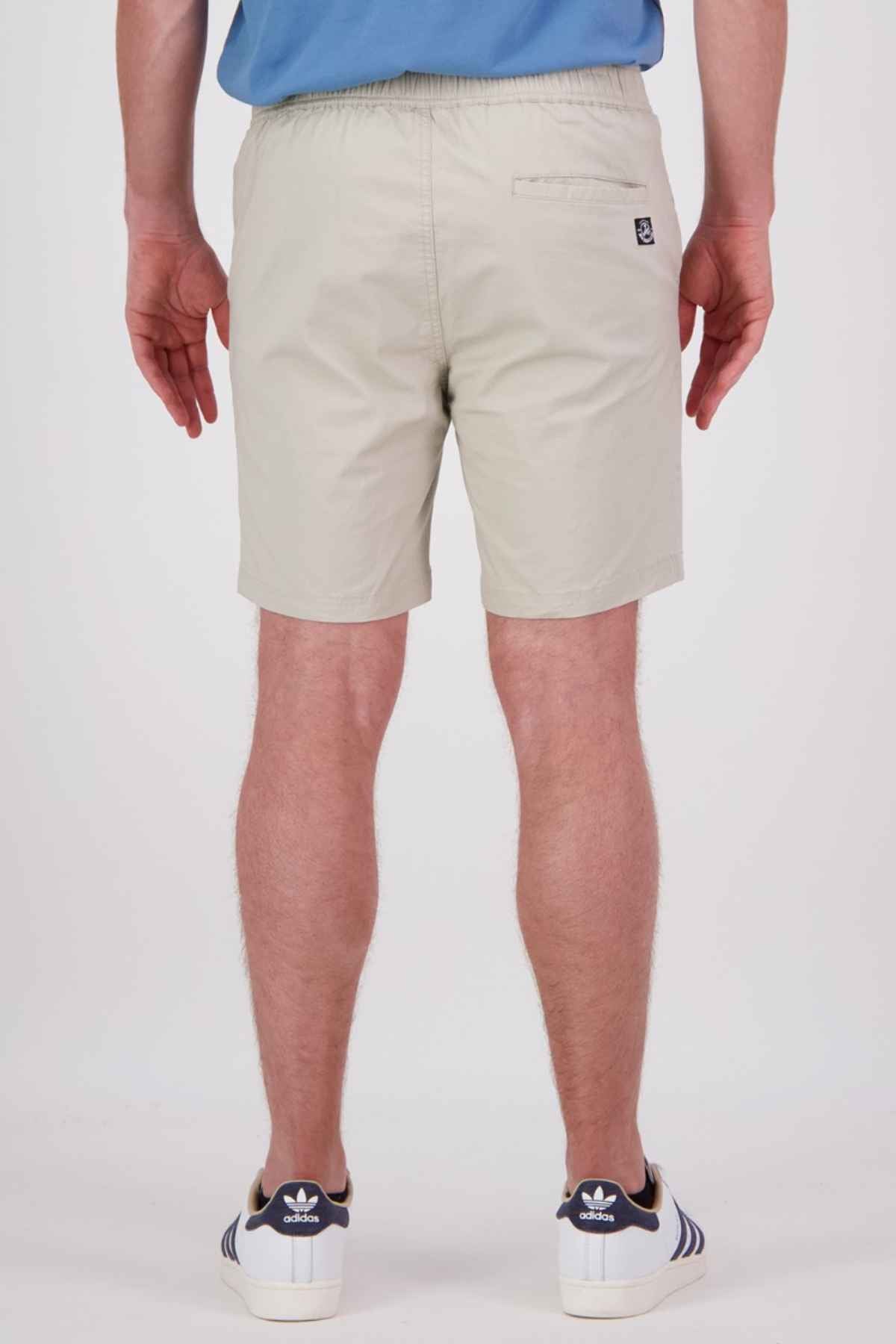 A person wearing Swanndri Sandy Point Shorts in light beige stretch cotton fabric, paired with a blue shirt and white sneakers featuring dark blue stripes, stands with their back to the camera. The shorts are designed with a hidden zip back patch pocket for added convenience.