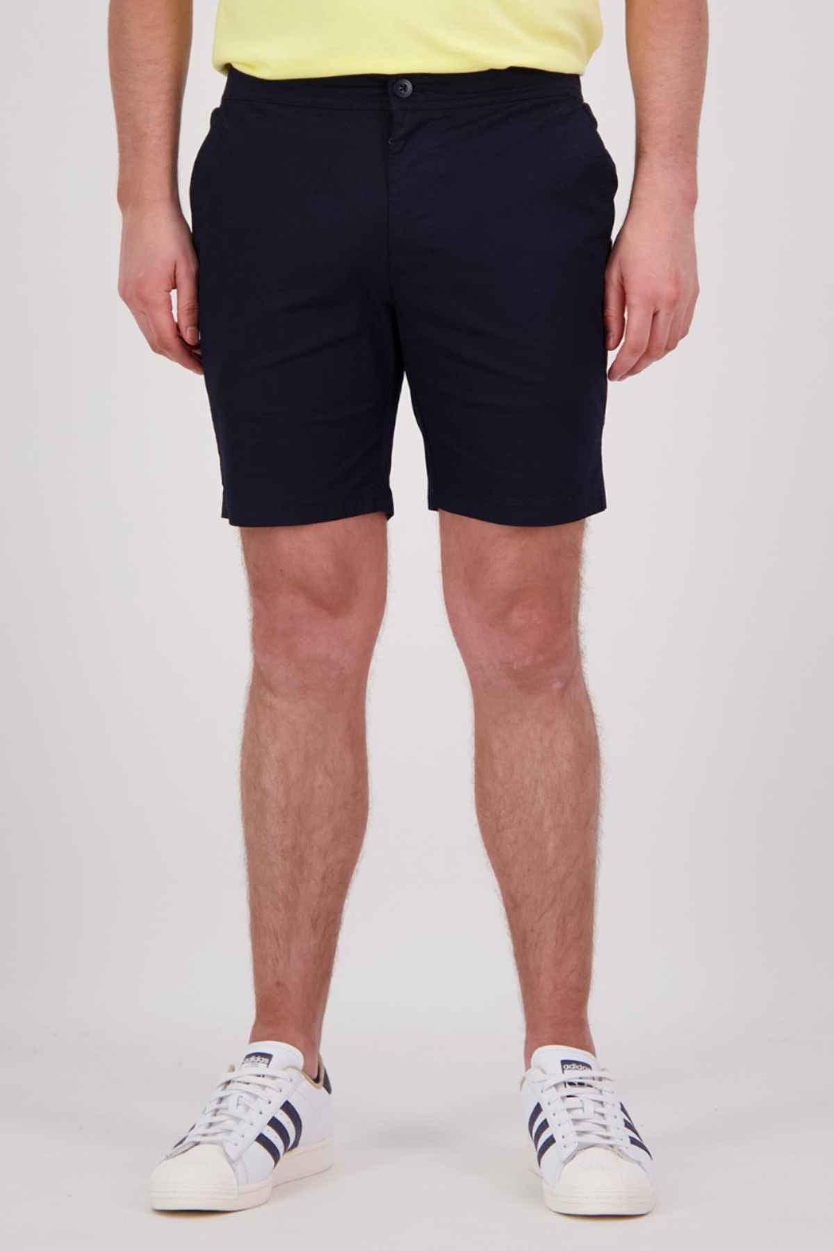 A person wearing navy blue Swanndri Sandy Point Shorts, crafted from a cotton stretch fabric, and white sneakers with black stripes stands in front of a plain background. While their upper body is not shown, the shorts are highlighted by their multiple pockets for added versatility.