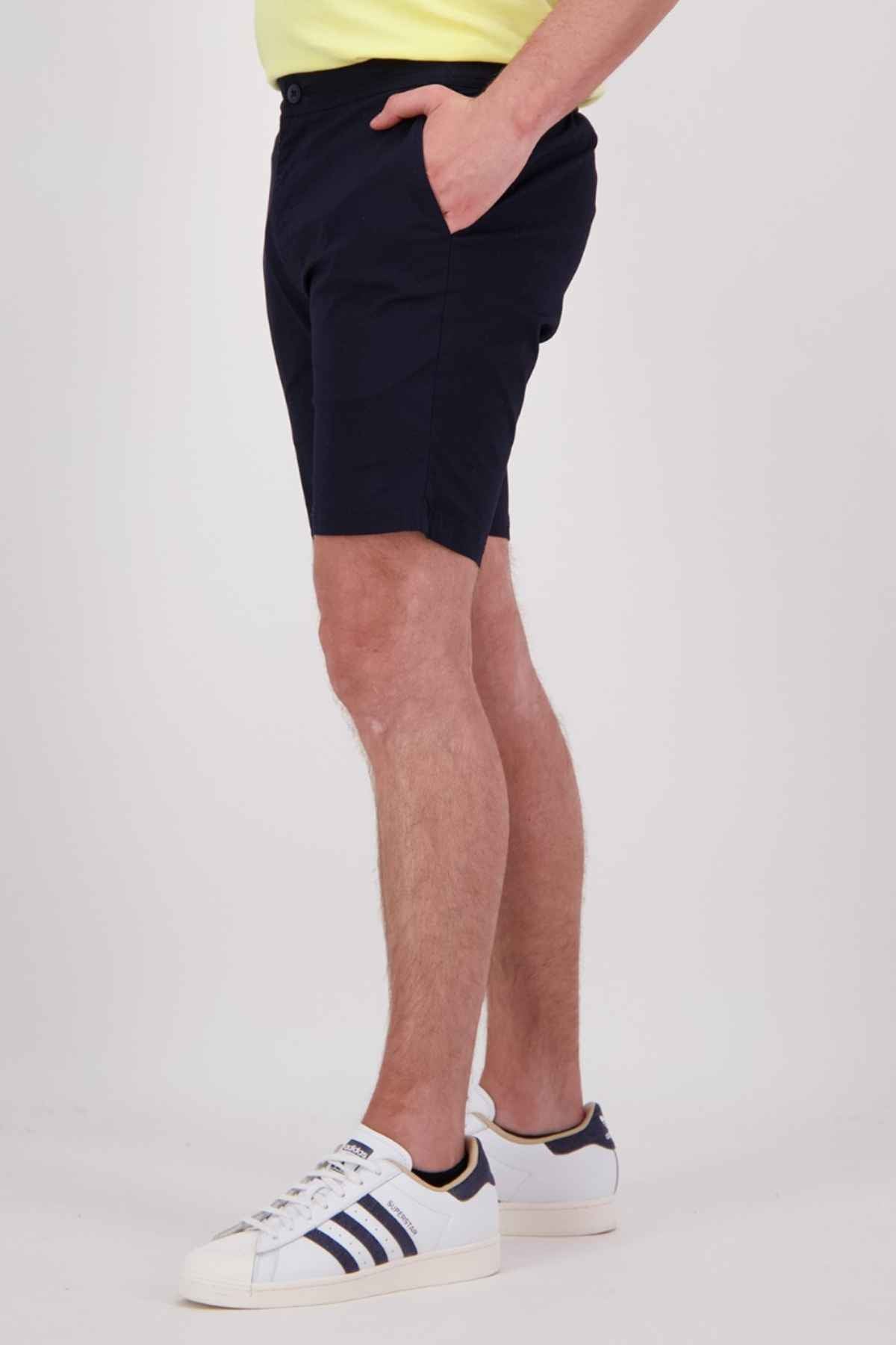 A person is standing against a plain background wearing Swanndri Sandy Point Shorts in navy blue, crafted from cotton stretch fabric. They have paired these shorts with a light-colored shirt and white sneakers that feature blue stripes. One of their hands rests in one of the multiple pockets of the shorts.