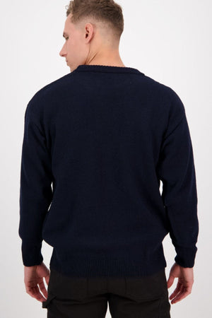 A man with short hair stands with his back to the camera, wearing a Swanndri Seafarer Crew Neck Sweater in dark navy blue and black pants against a plain light background.