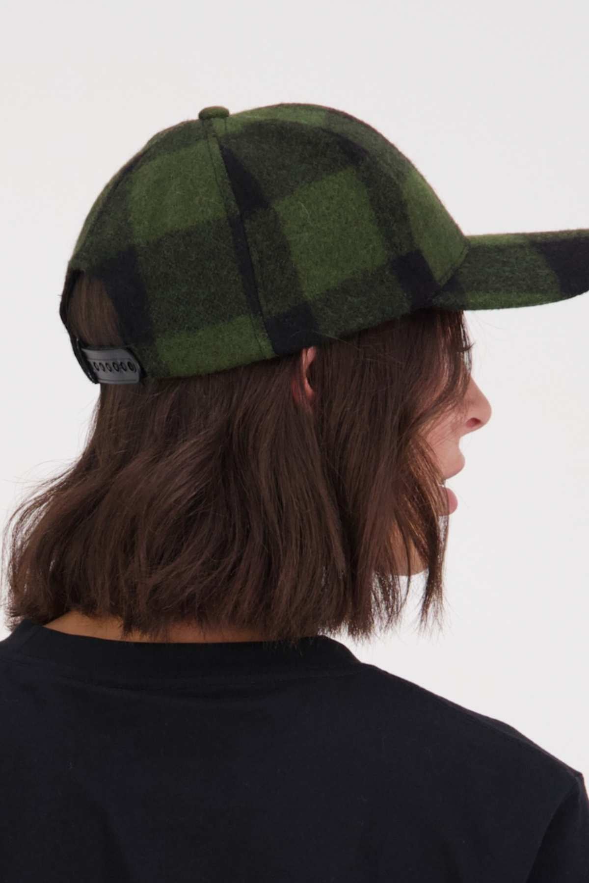 A person with shoulder-length brown hair wears a black shirt and a green and black Swanndri Swanni Wool Check Baseball Cap, shown from the back and side view, against a light background.