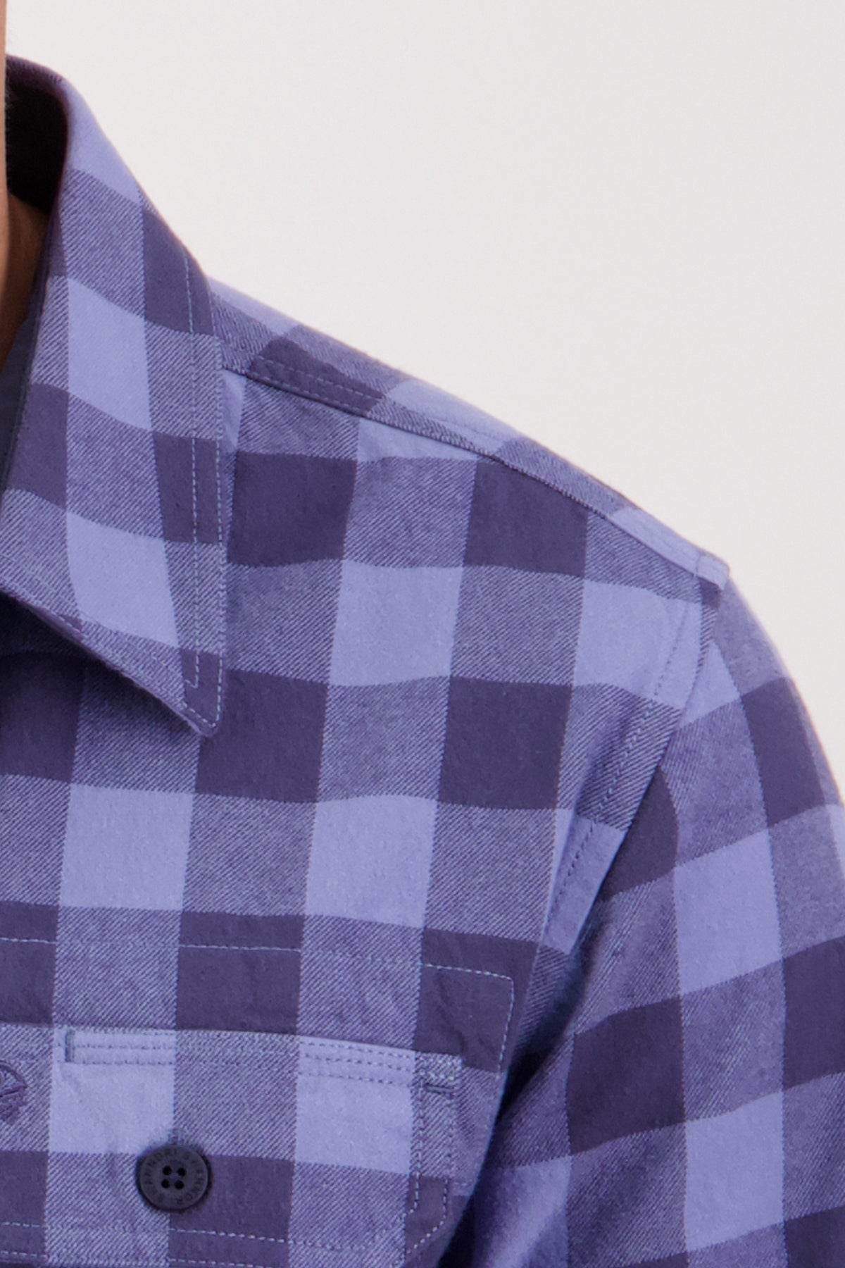 Close-up of someone wearing the Swanndri Taranaki Tailor Shirt, featuring a blue and dark blue checkered pattern, collar, and buttoned chest pockets; background is plain and light.