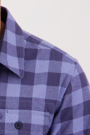 Close-up of someone wearing the Swanndri Taranaki Tailor Shirt, featuring a blue and dark blue checkered pattern, collar, and buttoned chest pockets; background is plain and light.