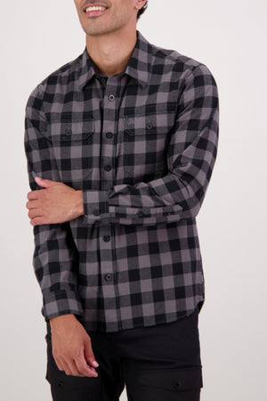 A person in a Swanndri Taranaki Tailor Shirt—a long-sleeve, black and gray men's casual shirt—stands against a plain light background with one arm crossed, smiling slightly.